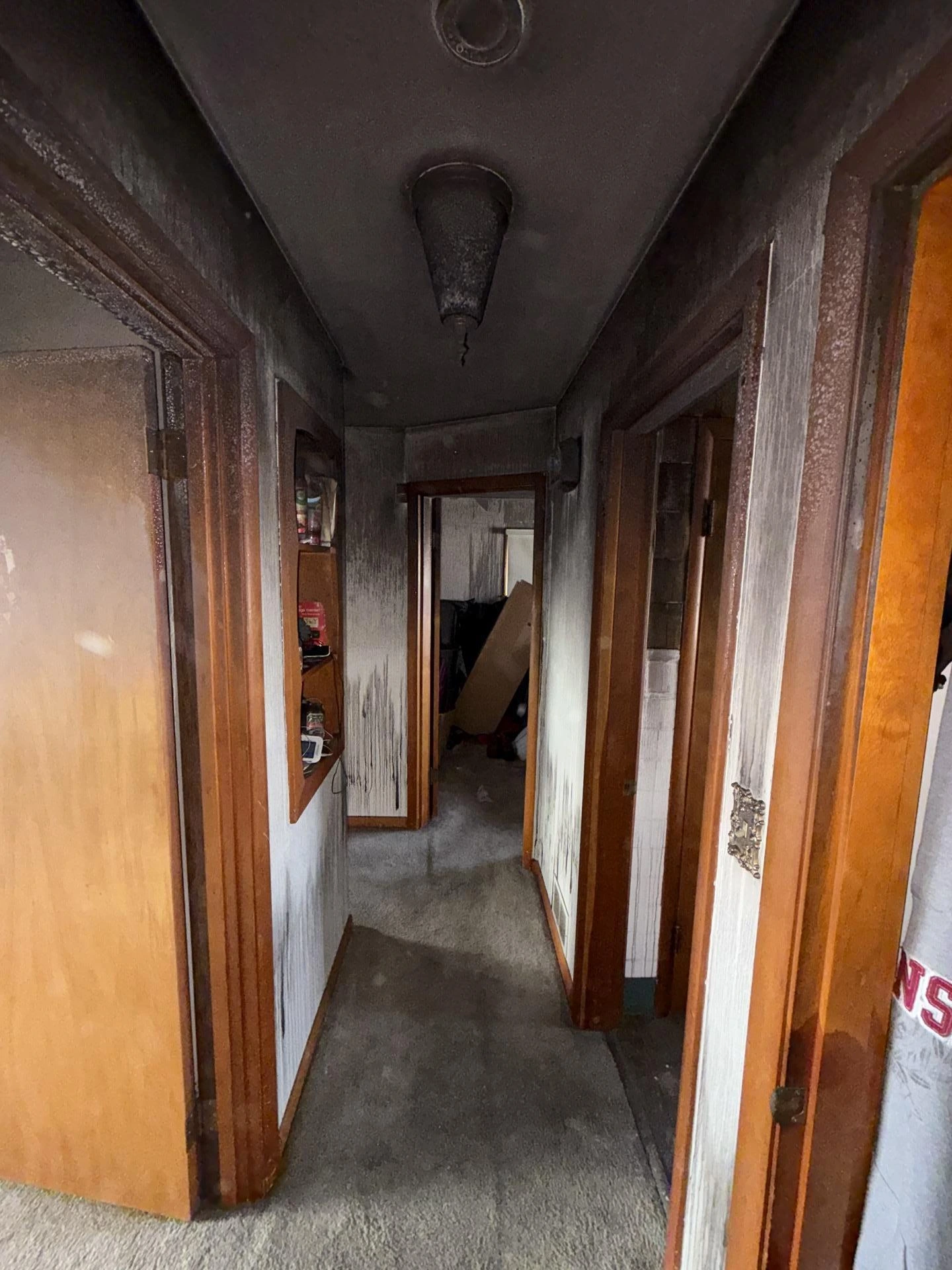 Residential hallway coated in heavy black soot and smoke damage after a house fire, with darkened ceiling, smoke-stained walls, scorched door frames, and debris visible in the room at the end of the corridor