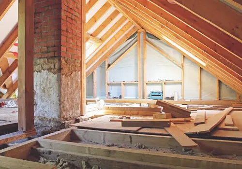 Residential attic under reconstruction showing exposed wooden roof rafters, brick chimney, insulation, and lumber materials during a home restoration or renovation project