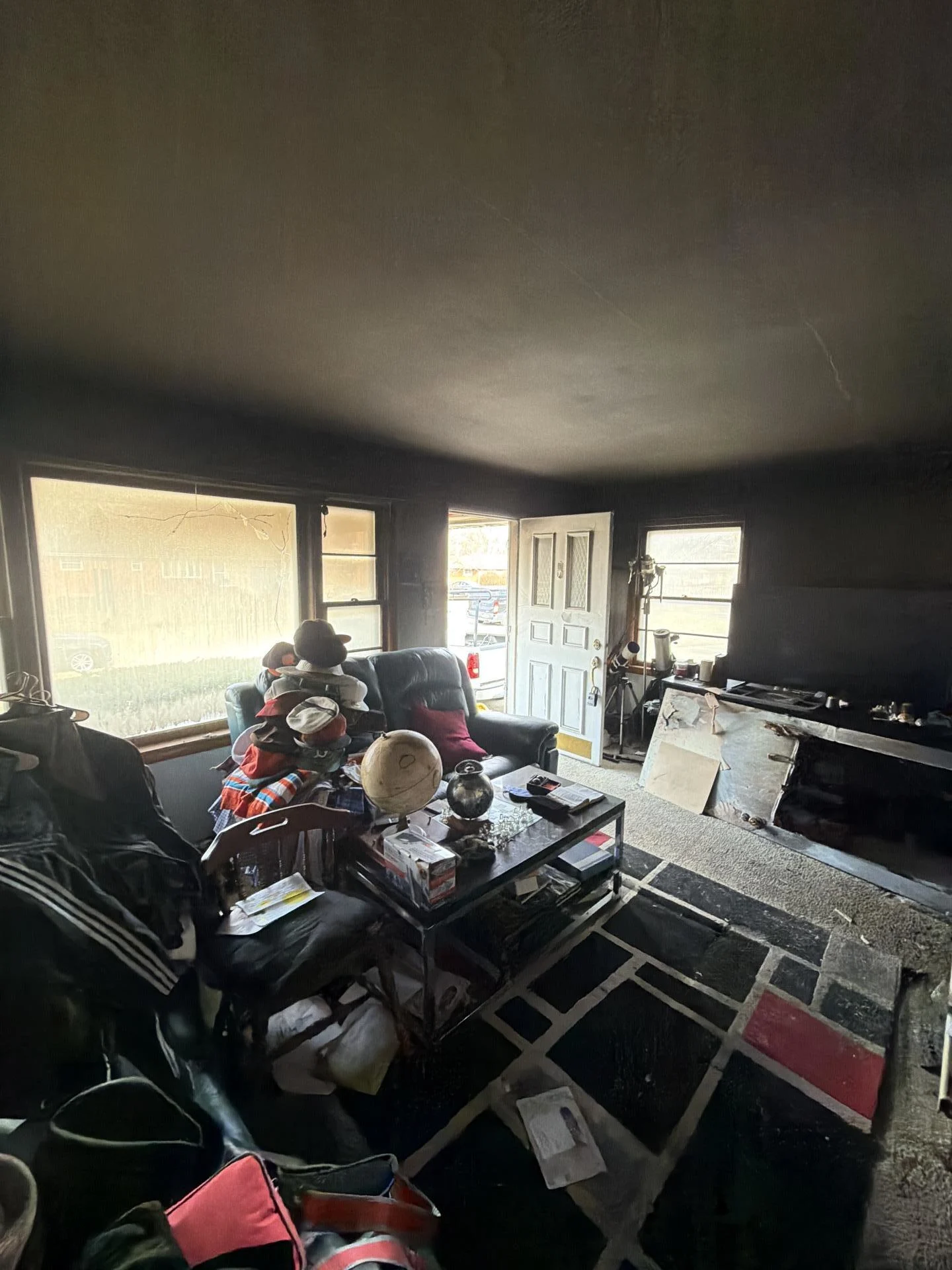 Smoke-damaged living room with soot-blackened ceiling and walls, ash-covered furniture and belongings, burned carpet, and smoke-filmed windows after a residential house fire