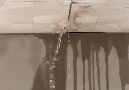 Water actively streaming through a cracked ceiling with visible water stains and moisture damage spreading across the surface, showing an active indoor water leak emergency