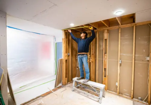 Contractor standing on a step stool measuring exposed wooden wall framing during an interior home reconstruction or water damage restoration rebuild