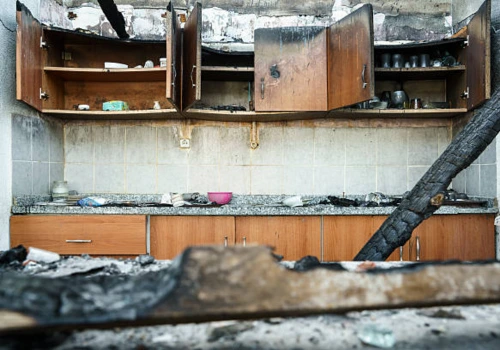 Severely fire-damaged kitchen with charred cabinets, burned ceiling, soot-covered countertops, and collapsed structural debris, showing extensive fire and smoke damage requiring full restoration
