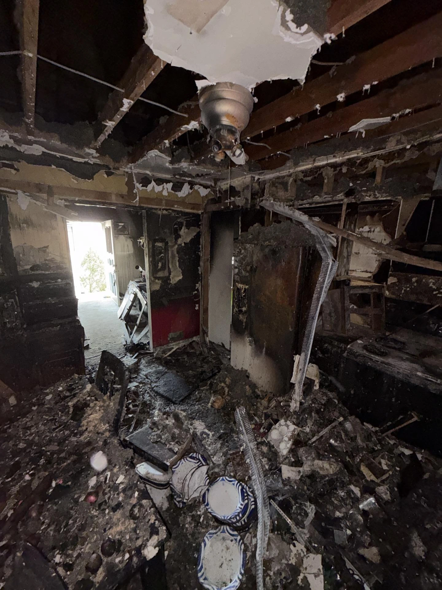 Completely destroyed home interior after a severe house fire, showing charred walls, collapsed ceiling with exposed burnt rafters, ash-covered debris on the floor, and a melted ceiling fan fixture