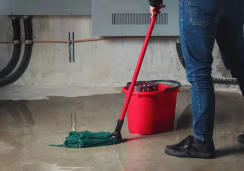 Person mopping a wet flooded basement floor with a red bucket beside them, showing water damage cleanup near exposed pipes and an electrical panel
