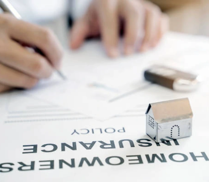 Person signing a homeowners insurance policy document with a miniature house model and key on the desk, representing property insurance coverage for water or fire damage claims