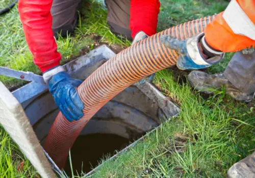 Two workers in protective gloves feeding a large industrial suction hose into an outdoor sewer or septic tank opening during a sewage cleanup or drain service operation