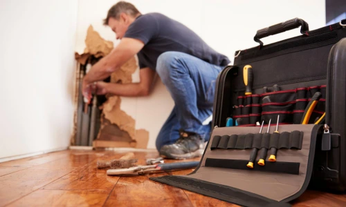 technician repairing water damaged wall with exposed pipes and tools on hardwood floor