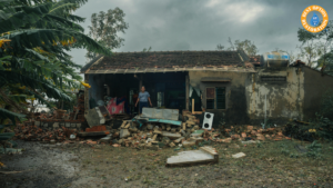 A person stands in front of a damaged house with a collapsed roof and scattered debris. The scene conveys a sense of devastation and loss.