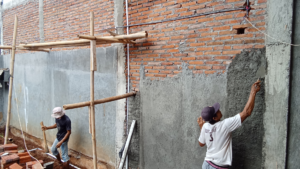 Two construction workers apply cement to a red brick wall. One smoothes the surface, while the other stands nearby, surrounded by bamboo scaffolding.