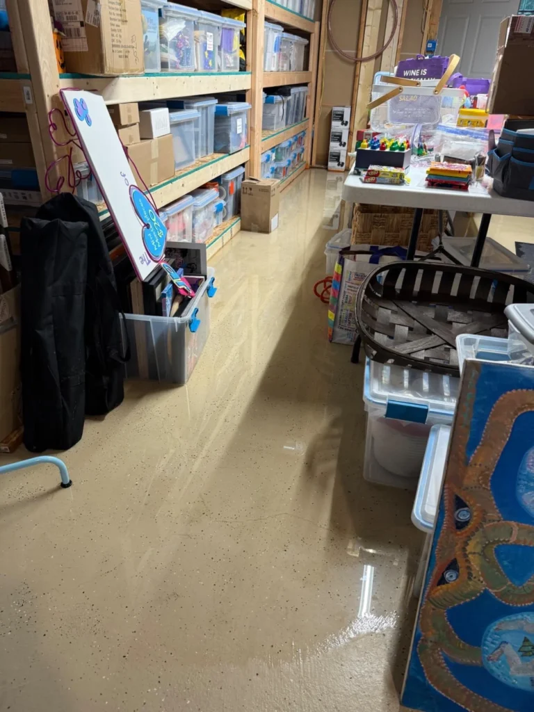 Flooded basement craft storage room with standing water on epoxy floor, wooden shelving filled with labeled plastic storage bins, folding table with supplies