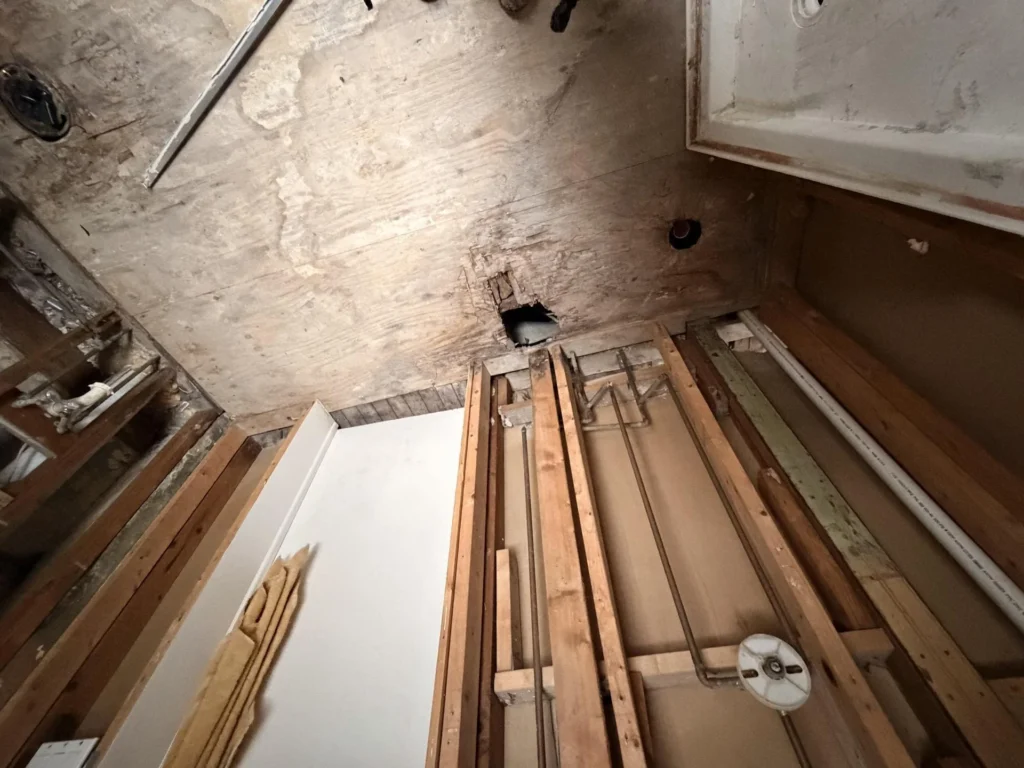 Upward view of bathroom showing damaged subfloor from above with hole, exposed wood framing and pipes, lumber stacked below during water damage restoration