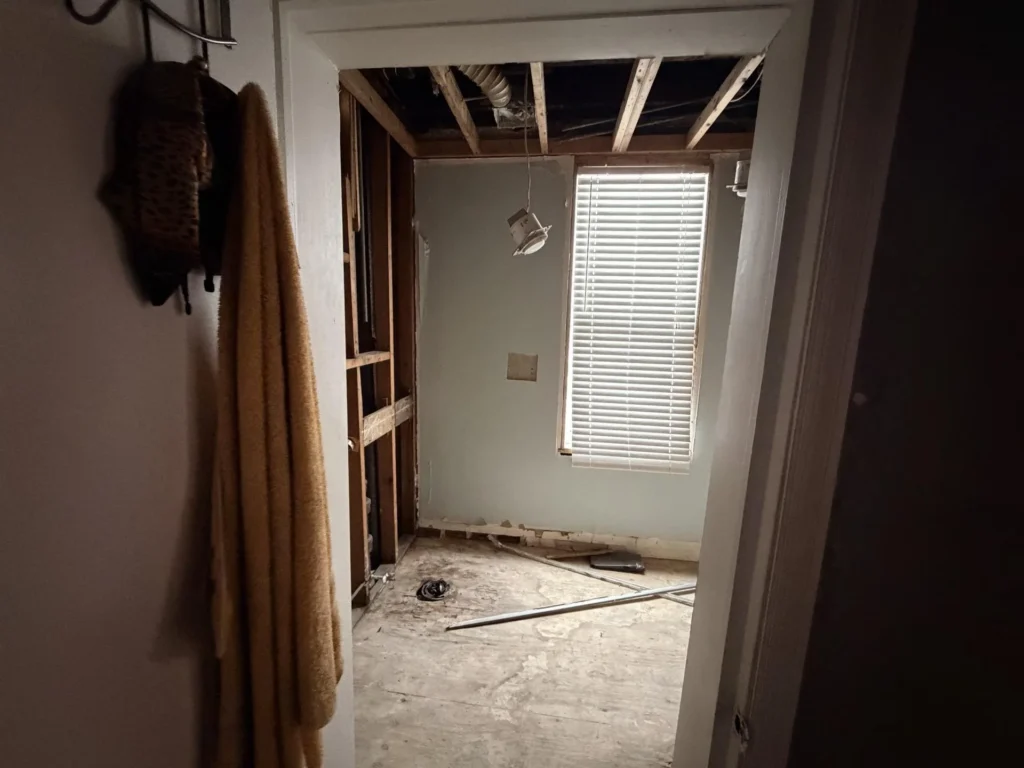 View through doorway into stripped bathroom showing exposed wood studs, dangling ceiling light fixture, window with blinds, bare subfloor with debris, coat hook with towel in foreground