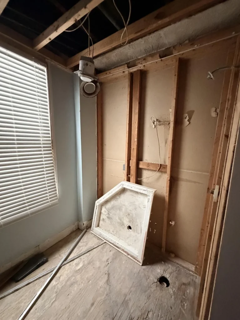 Bathroom stripped to studs with removed shower pan leaning against wall, open ceiling joists, window with blinds, bare subfloor with drain hole exposed