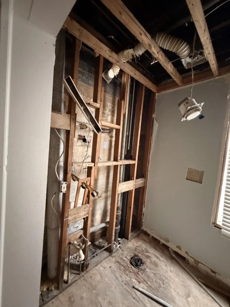 Bathroom gut-renovation showing exposed wood stud wall with plumbing and electrical, arched ceiling opening, dangling light fixture, window with blinds on far wall