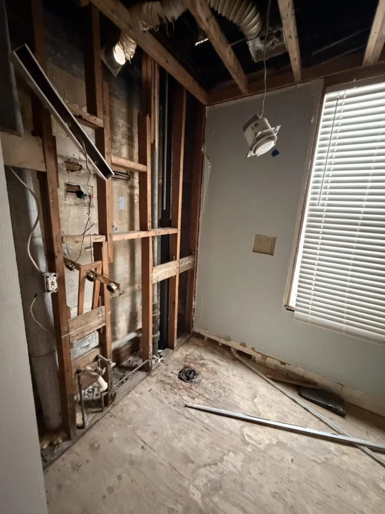 Bathroom stripped to wood studs with exposed plumbing rough-in, HVAC flex duct visible at open ceiling, window with blinds intact, bare subfloor with debris