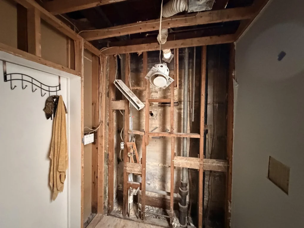 Bathroom wall fully stripped to wood studs exposing plumbing pipes, electrical conduit, and HVAC ductwork after water damage remediation, coat rack with towel on adjacent wall