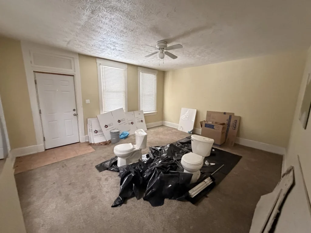 Bedroom used as staging area with two removed toilets sitting on black plastic sheeting, drywall debris, moving boxes, and construction materials on carpeted floor
