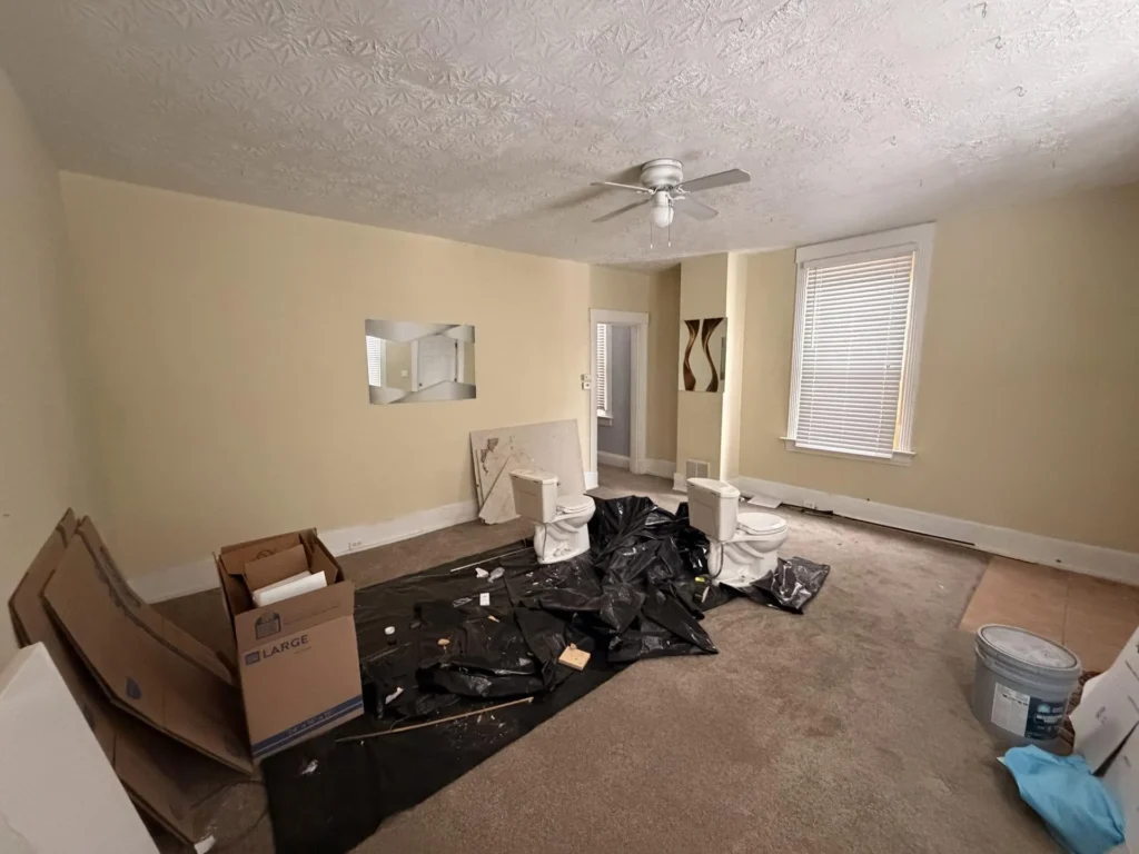 Wide view of bedroom with two removed toilets on black plastic sheeting, Content Restorology boxes, moving cartons, ceiling fan, textured ceiling with water stains, exterior door
