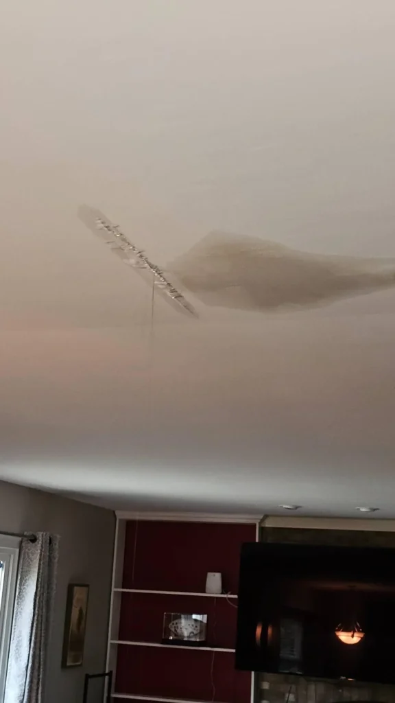 Living room ceiling showing long diagonal crack with separated drywall and large water stain bulge, red accent wall with TV and shelving visible below