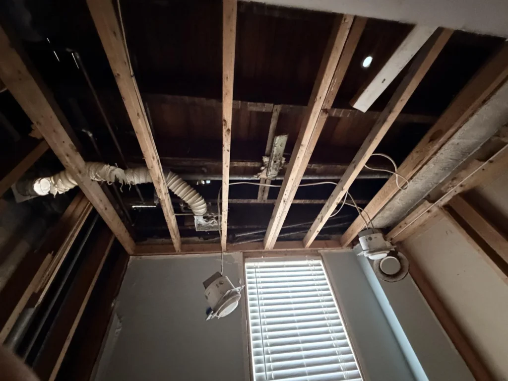 Looking up at fully exposed ceiling with wood joists, flexible HVAC duct, electrical wiring, and dangling light fixtures after ceiling drywall removal due to water damage