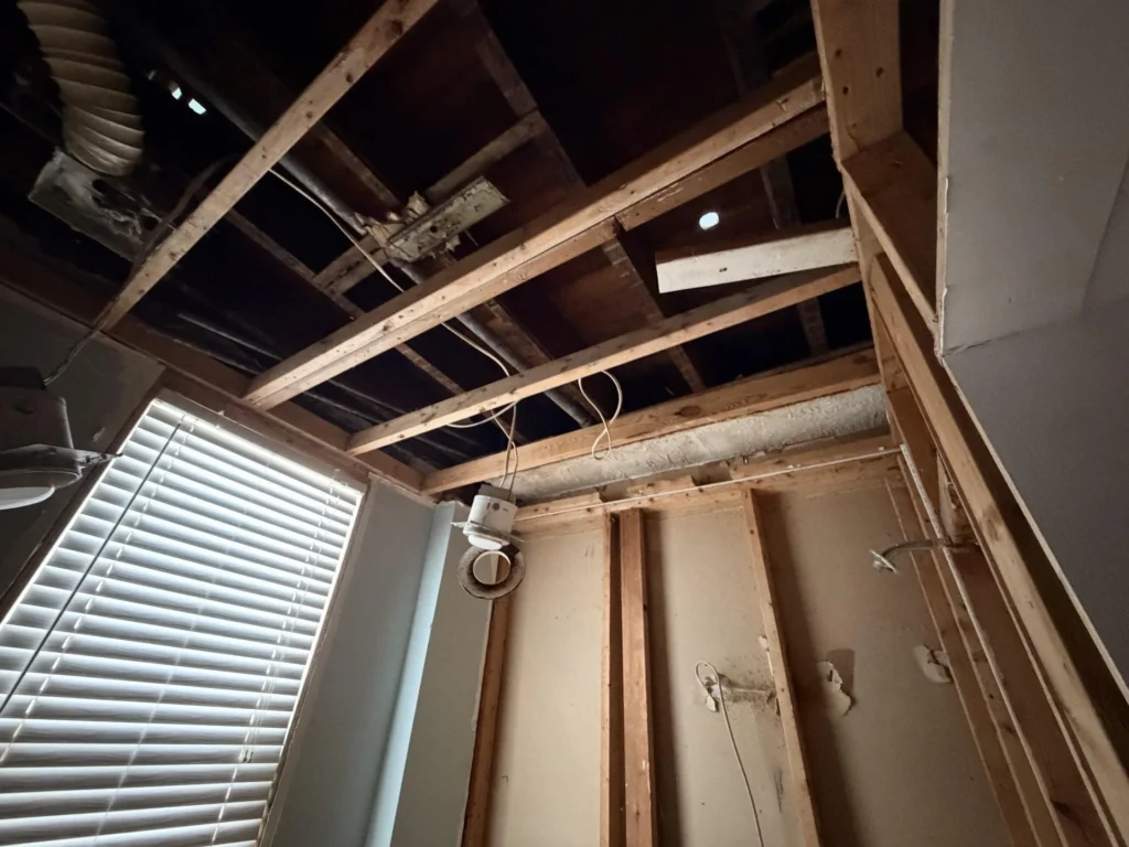 Angled upward view of stripped ceiling showing wood joists, wiring, and open ceiling cavity with window blinds visible below and partial drywall remaining on wall