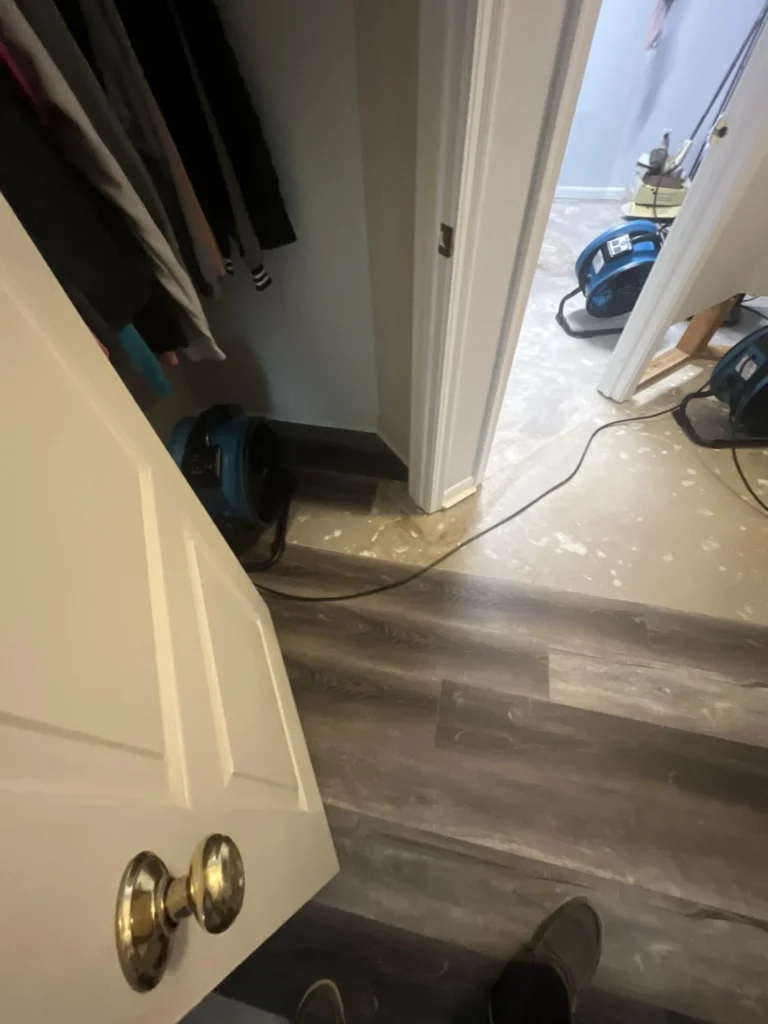 View from closet doorway showing blue air mover fans deployed on bare concrete subfloor for drying during water damage remediation