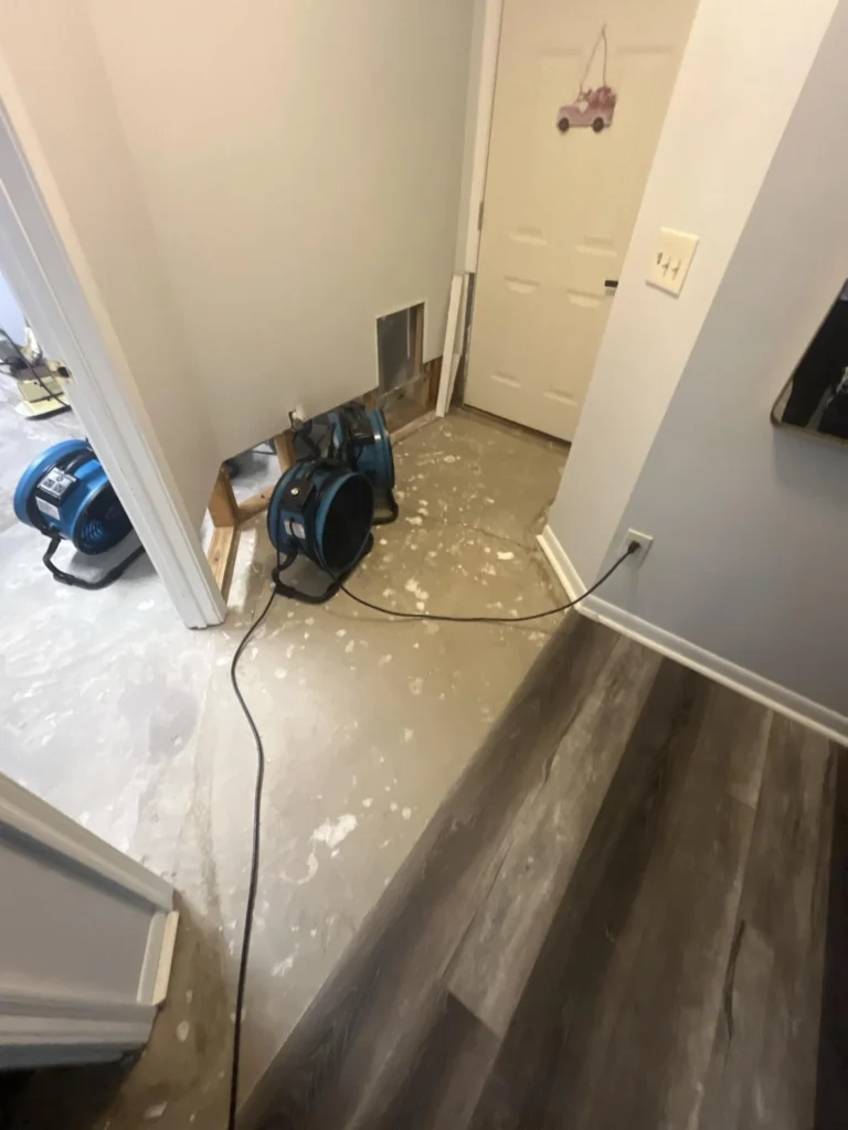Entryway and hallway with bare concrete subfloor and blue air mover fans positioned for drying, wall cutout for remediation, front door visible
