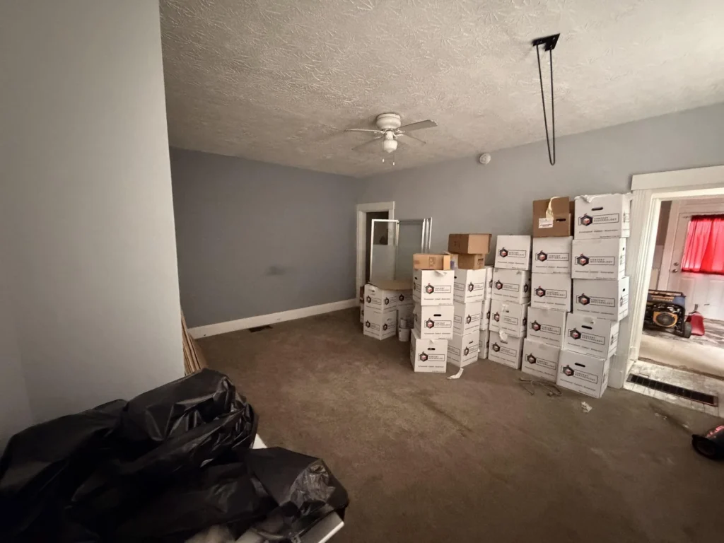 Entry area looking down staircase toward door, Content Restorology moving boxes stacked, Firman generator visible in kitchen, stripped subfloor with floor register, shuttered window
