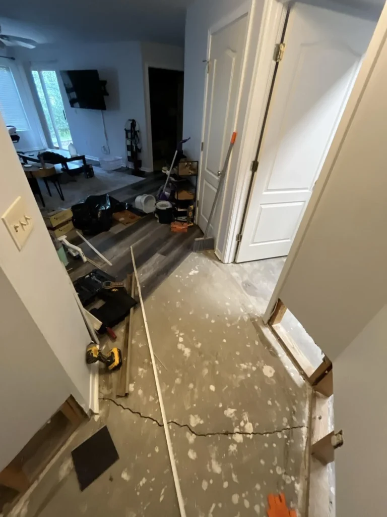 Hallway with all flooring removed exposing bare concrete subfloor, power tools and debris scattered, living room with intact flooring visible beyond
