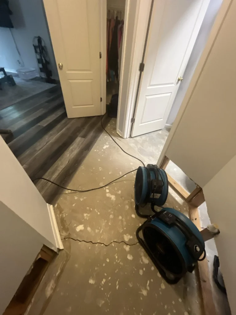 Hallway with two blue air mover fans positioned on bare concrete subfloor for drying, exposed wall framing visible, doors to closet and bedroom open