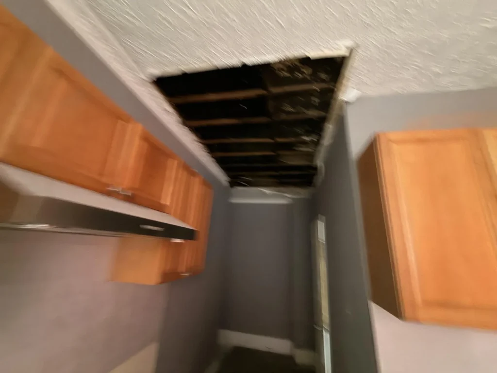Kitchen with large hole in textured ceiling exposing dark joists above, honey oak upper cabinets intact, range hood visible, gray walls during water damage remediation