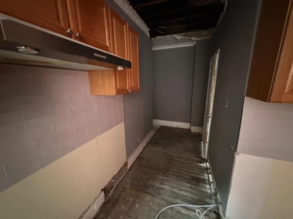 Narrow kitchen corridor with honey oak upper cabinets, range hood, gray subway tile backsplash partially removed, open ceiling exposing black joists, damaged wood floor