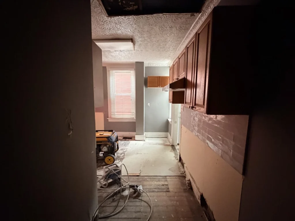 Water-damaged kitchen viewed from entry, stripped subfloor with wood planks exposed, upper cabinets remaining, drywall removed at lower walls, generator and hoses on floor, ceiling partially open showing joists