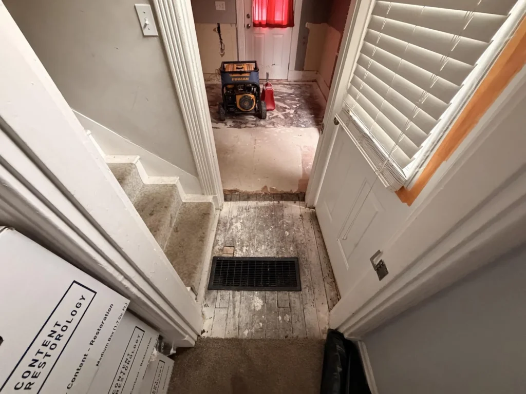 Wide view of water-damaged kitchen showing Firman generator, honey oak upper cabinets with range hood, stripped lower walls, bare subfloor, window with red curtain and exterior door