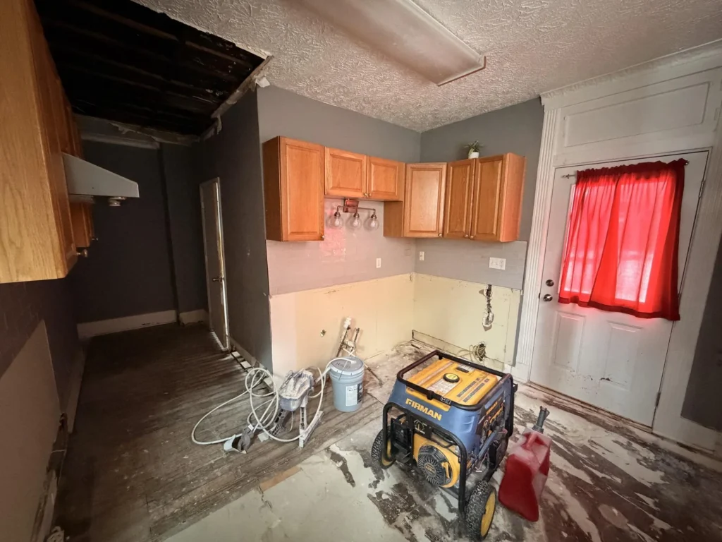 Kitchen during remediation with Firman generator and fuel can, upper cabinets remaining, lower drywall removed, bare subfloor, red curtain on window near exterior door, ceiling hole visible