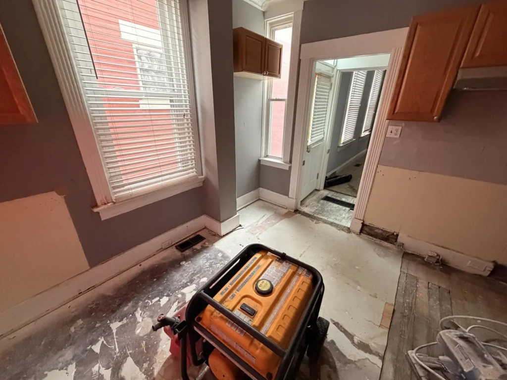 Kitchen with stripped subfloor, orange Firman generator on floor, tall windows with blinds, remaining upper cabinets, drywall removed at lower walls, doorway to adjacent room visible