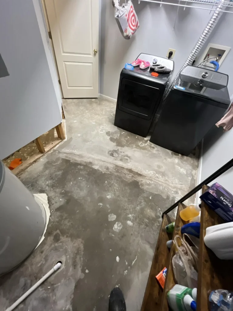 Laundry room with flooring completely removed exposing bare concrete subfloor, black washer and dryer, cut-out in lower wall for remediation