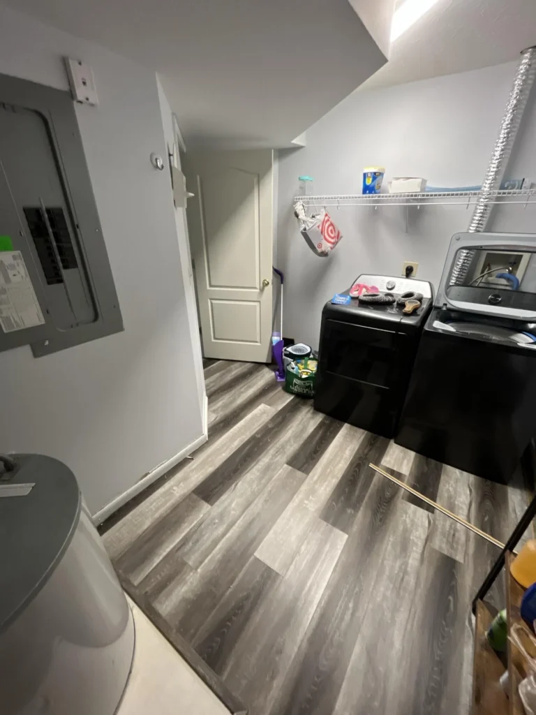 Laundry room with new gray luxury vinyl plank flooring installed, black washer and dryer units, electrical panel and wire shelving visible