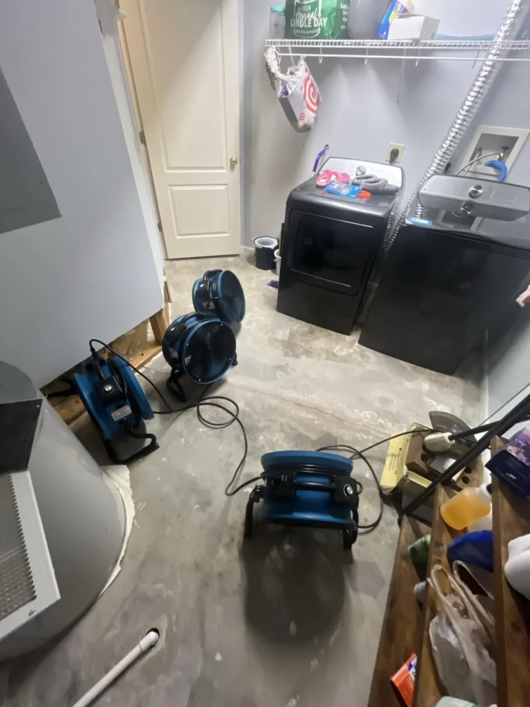 Laundry room with three blue air mover fans on bare concrete subfloor during remediation, black washer and dryer units, exposed wall framing