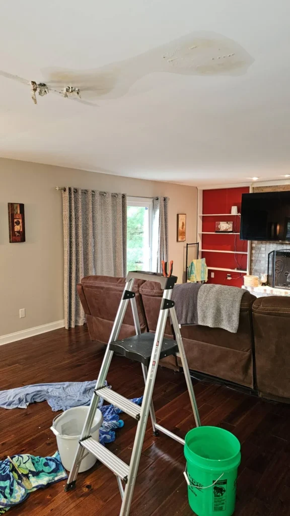 Living room ceiling with large water-damaged bulging drywall area and torn section, stepladder and collection buckets on hardwood floor below