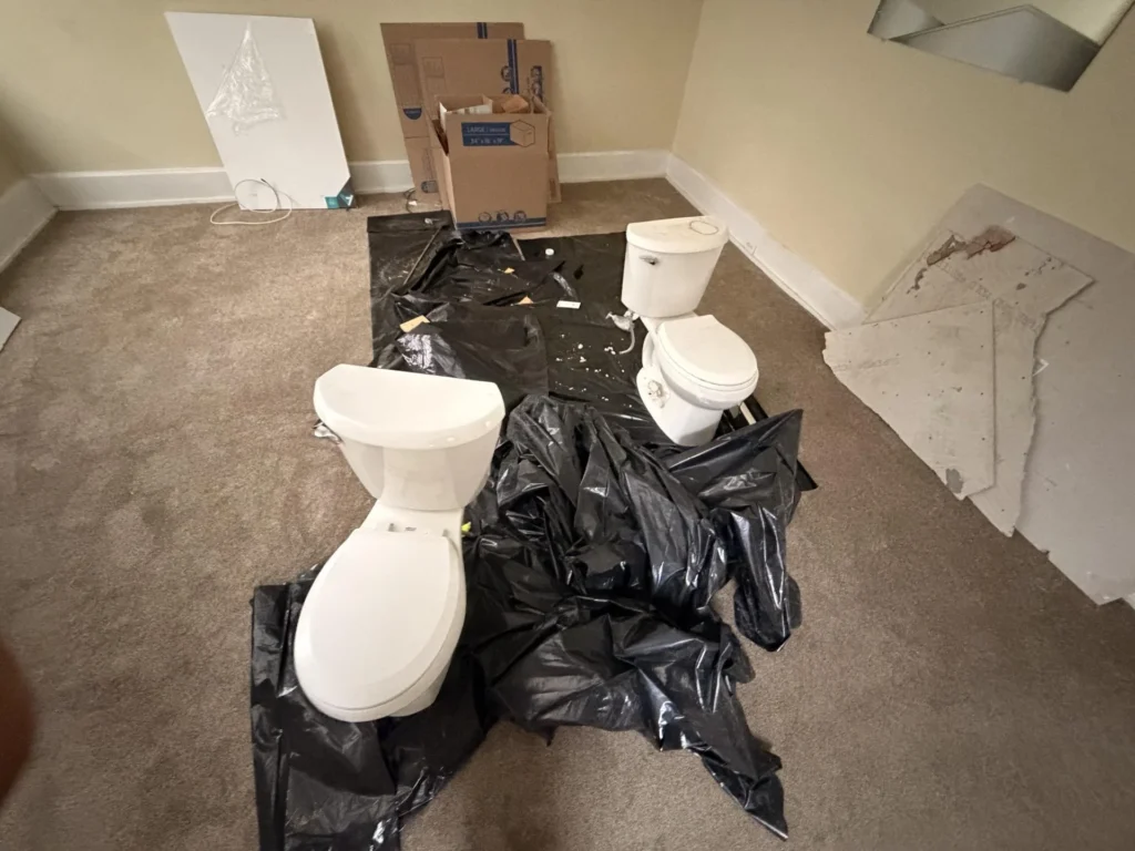 Living room with carpet intact, stacked Content Restorology restoration boxes along wall, black trash bags on floor, ceiling fan, light fixture wire hanging, doorway to kitchen visible