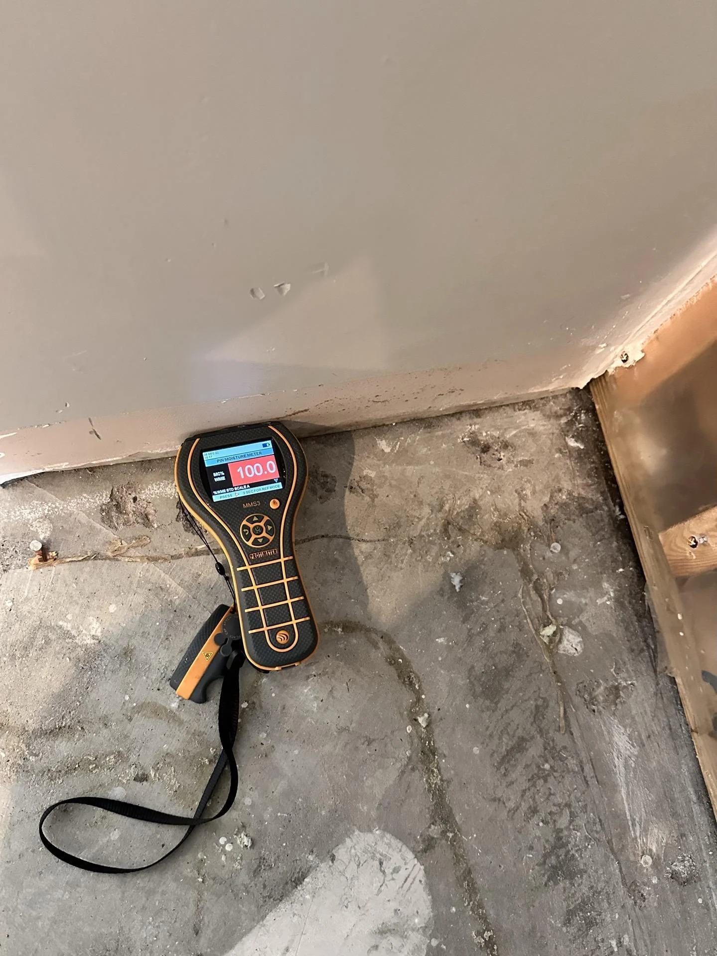 Moisture meter showing reading of 100.0 percent placed at baseboard level on concrete subfloor near water-damaged drywall