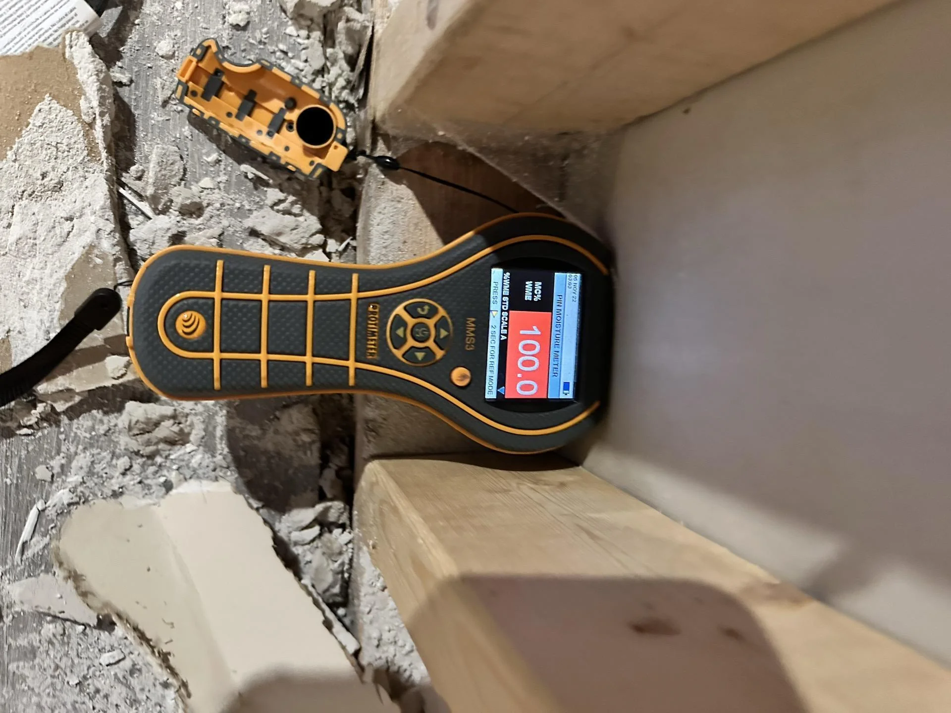 Moisture meter (Orlihle MMS3) displaying reading of 100.0 percent inserted into opened wall cavity with exposed lumber framing and concrete debris