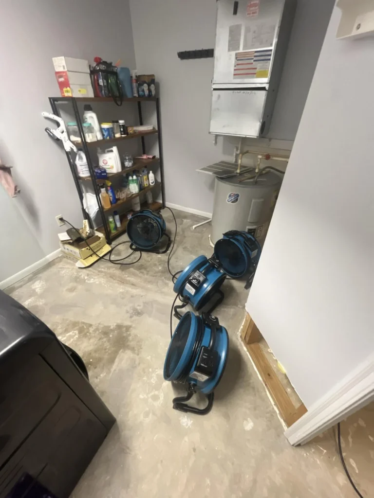 Utility room with four blue air mover fans deployed on bare concrete subfloor, water heater, furnace, and supply shelving in background
