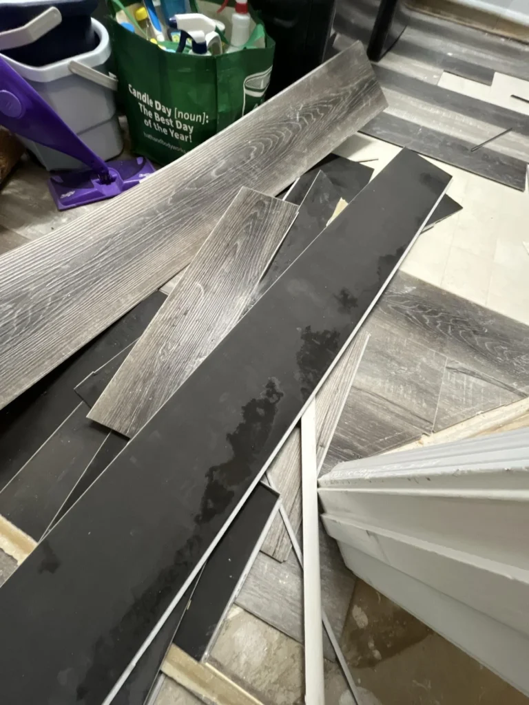 Pile of removed water-damaged luxury vinyl plank flooring with black backing residue and adhesive, stacked on remaining tile floor during remediation