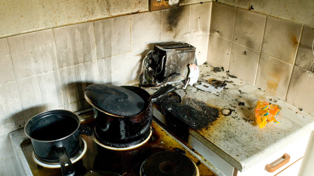 Charred kitchen with a burnt stove, blackened pots, and a destroyed toaster. Soot marks on tiles convey the aftermath of a fire, evoking distress.