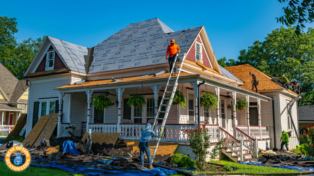 Workers in bright safety gear are repairing the roof of a charming Victorian-style house. Tarps and tools cover the yard, suggesting an active renovation.